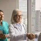 Smiling senior doctor shaking hands with a colleague while another medical professional looks on in a hospital office.