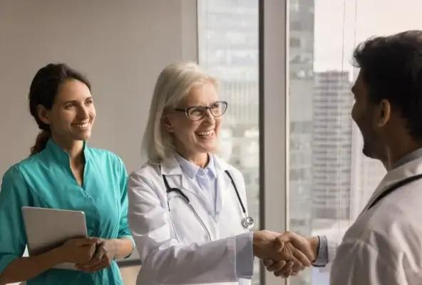 Smiling senior doctor shaking hands with a colleague while another medical professional looks on in a hospital office.