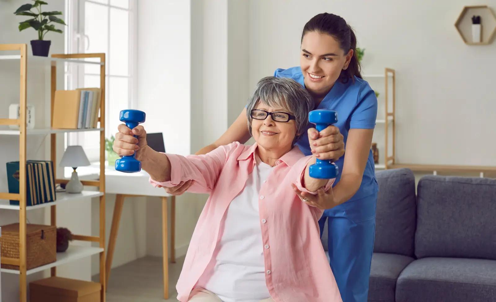 A physical therapist helps an older woman lift blue dumbbells during a rehabilitation or strength-training session in a bright home-like setting.